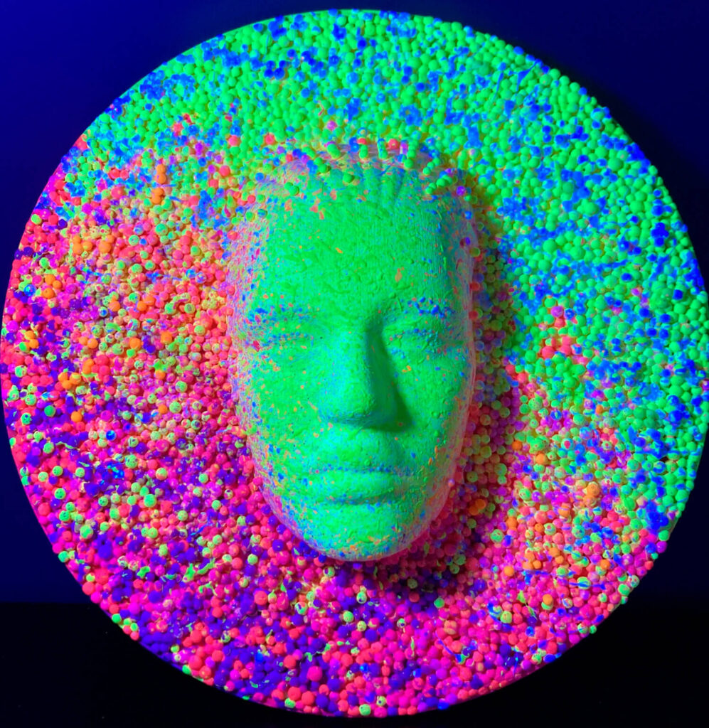 High-relief masks depicting a full face, rendered in a palette of yellow and shocking pink, with a pointillism effect created by small spheres forming the artwork